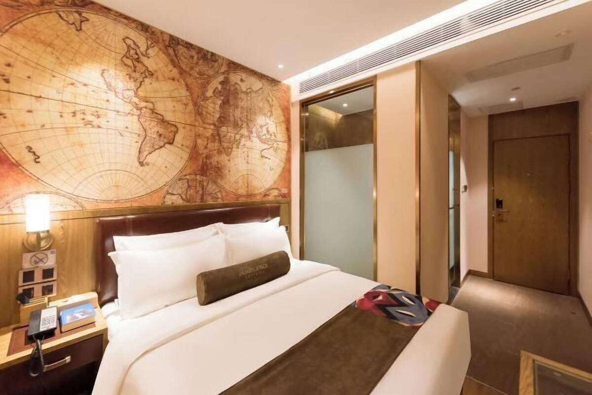 Habitación de hotel con un gran mapa mundial en la pared, una cama con ropa blanca, un cojín marrón y una mesita de noche con un teléfono y una lámpara.