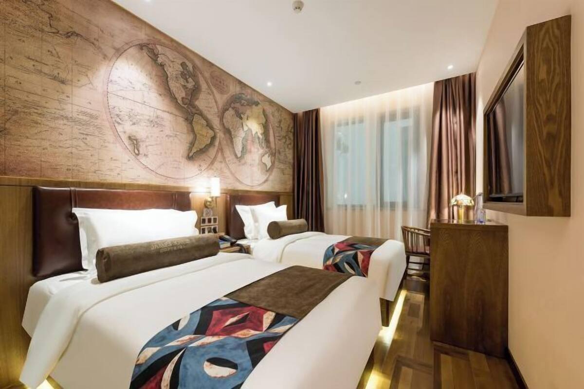 Habitación de hotel con dos camas, un gran mapa mundial en la pared, un escritorio con lámpara y una ventana con cortinas.