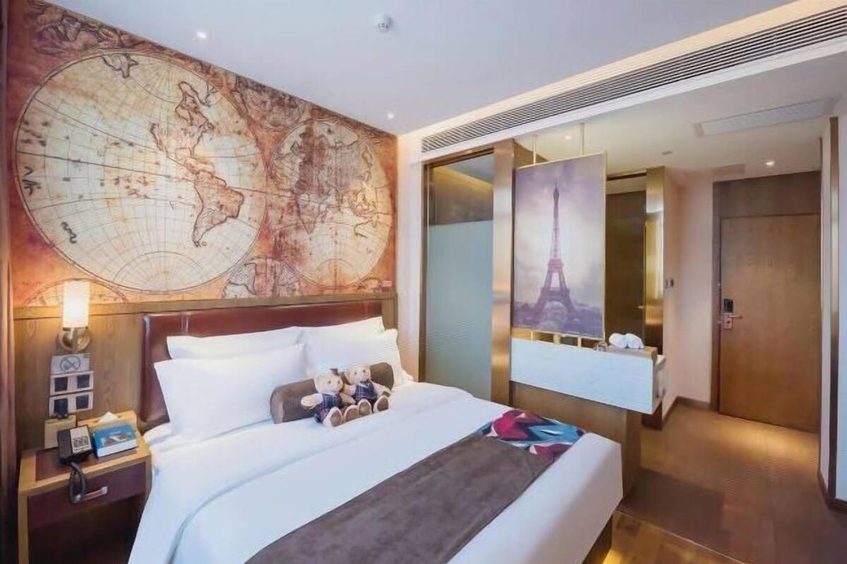 Habitación de hotel con un gran mapa mundial en la pared, una cama con ropa blanca, una mesita de noche con una lámpara y un cartel del Torre Eiffel.