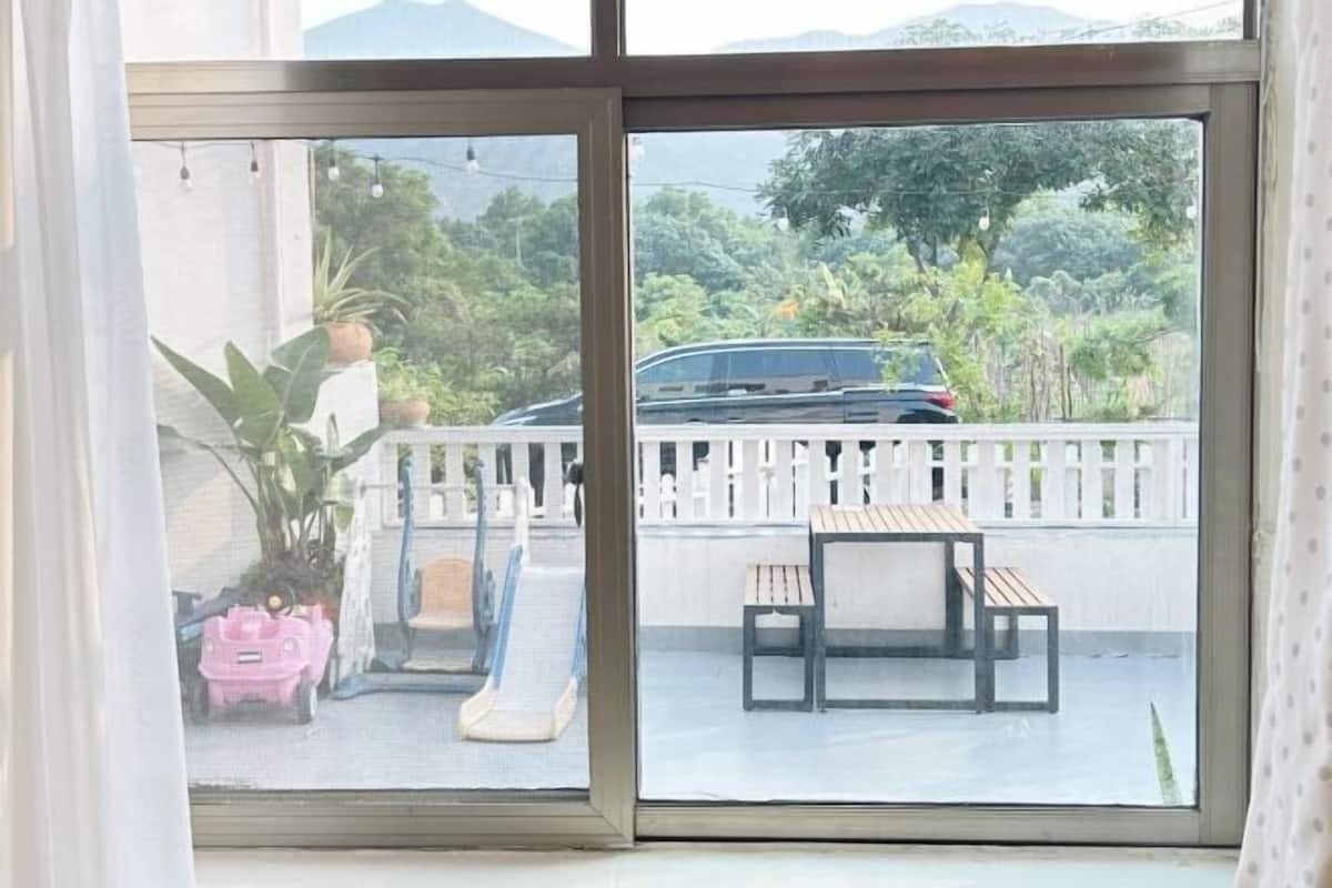 A view through a window with a balcony, featuring a car, a white fence, and greenery.