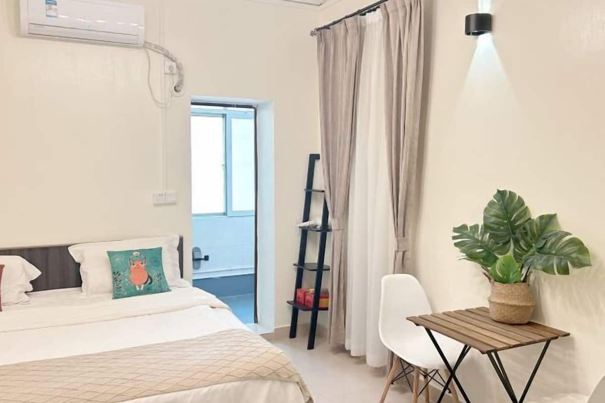 A bedroom with a bed, a small table with a plant, a ladder, and a view of the outside.