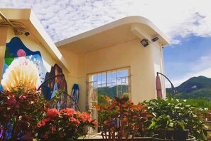 Un bâtiment orné d’une grande fresque sur le côté gauche, un balcon avec des plantes en pot et, au loin, une vue sur des montagnes.
