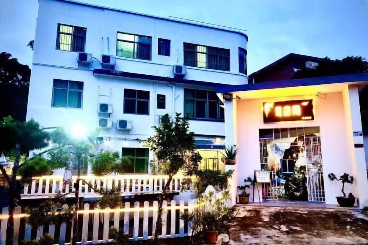 A two-story building with multiple windows and air conditioning units. There is a white fence in the foreground, and several potted plants are visible.