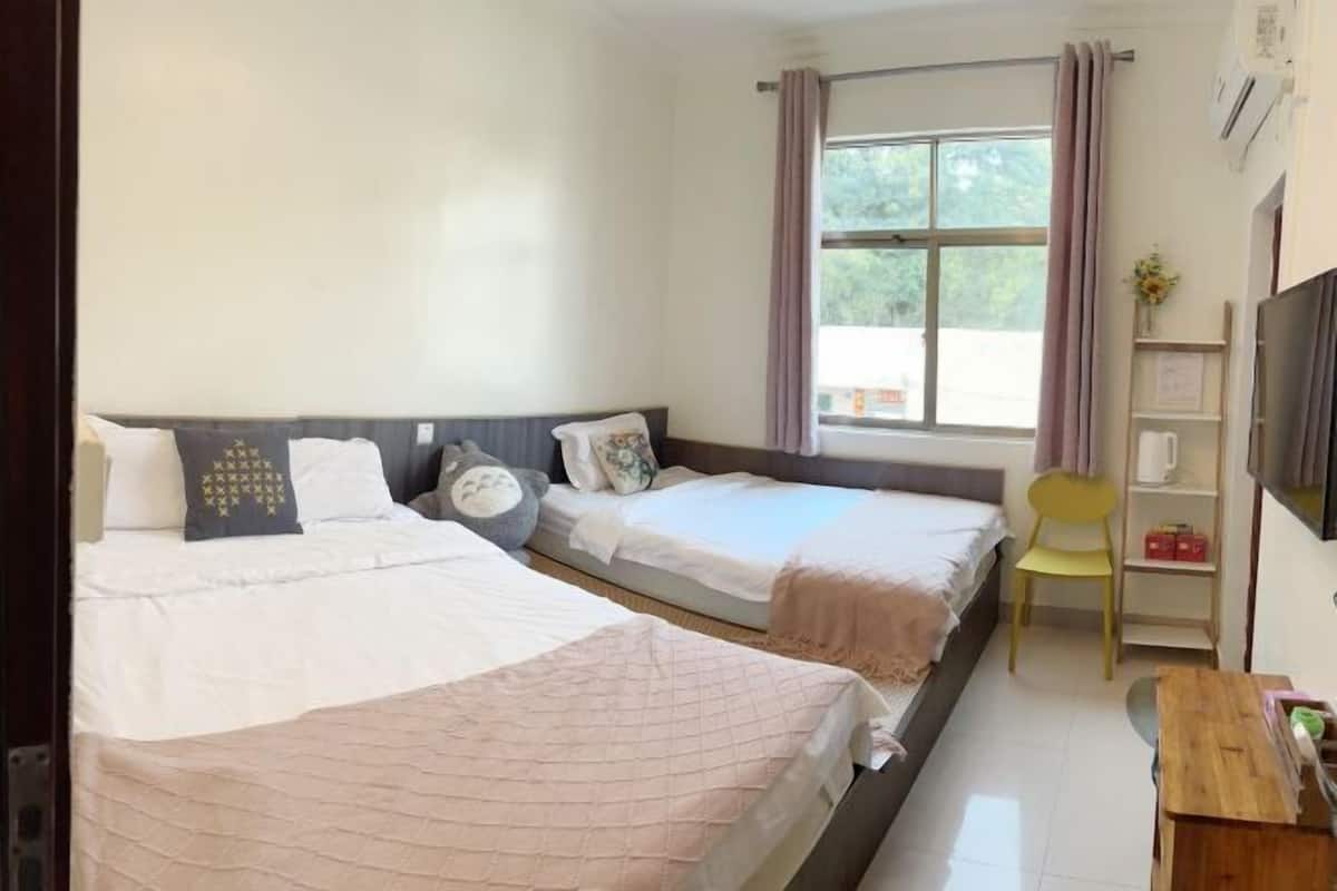 A room with two beds, a desk, a chair, a window with curtains, and a shelf with items.