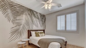 Une chambre à coucher avec un lit, un ventilateur de plafond, une fenêtre avec des stores, une petite table ronde et un mur orné d’un motif de feuilles.
