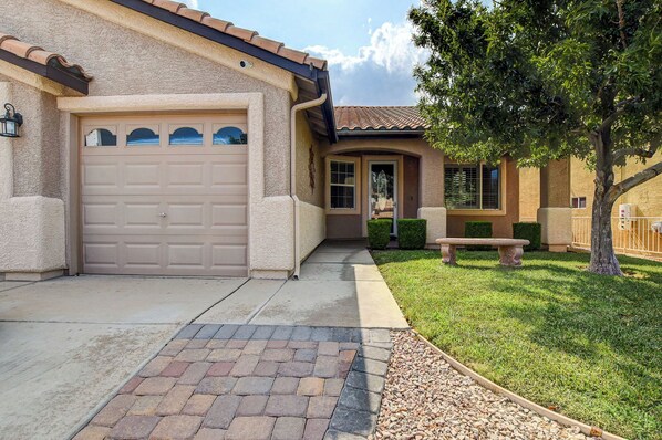 A residential house with a beige garage door, a front entrance, a small patio with a bench, and a well-maintained lawn.