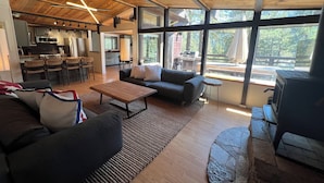 Amplia sala de estar con cocina moderna, chimenea de piedra y ventanas grandes con vistas al exterior.