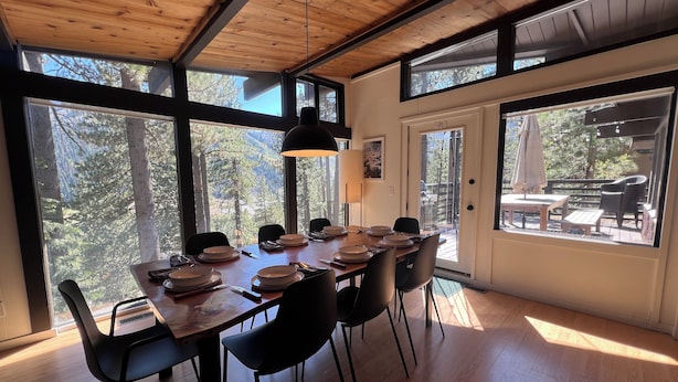 Comedor con mesa de madera dispuesta para seis personas, sillas negras, ventanas amplias con vistas a árboles y techo con vigas a la vista.