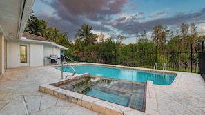 A swimming pool with a tiled deck, a small hot tub, and a covered area with a grill.