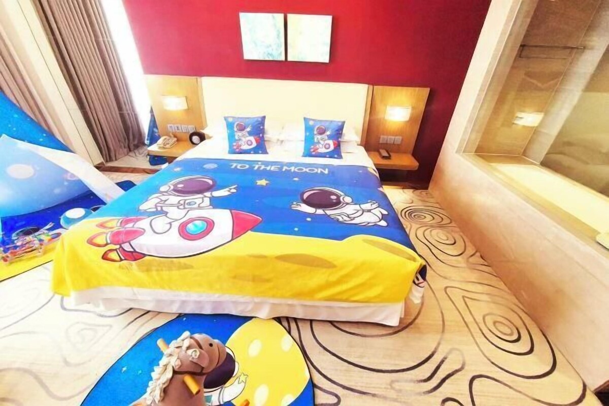 Una camera d'albergo con un letto, una coperta e dei cuscini a tema spaziale, un pavimento in legno con motivo circolare, una parete di colore rosso e una finestra con vista.