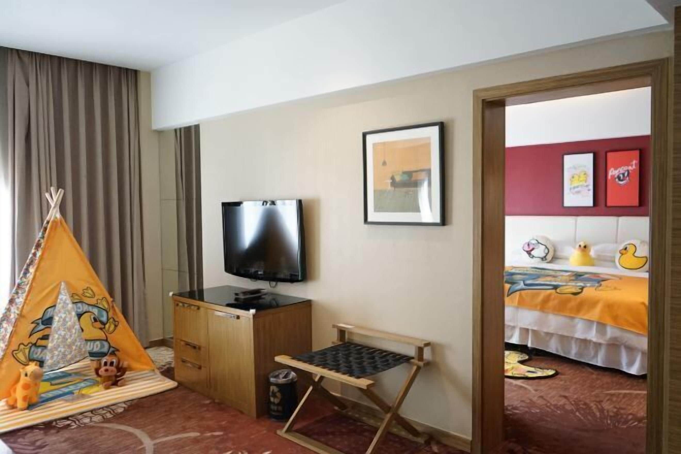 Habitación de hotel con televisión, una cama con colcha colorida, un escritorio pequeño con silla, una ventana con cortinas y una puerta que da a otra habitación.