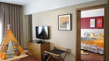 Habitación de hotel con televisión, una cama con colcha colorida, un escritorio pequeño con silla, una ventana con cortinas y una puerta que da a otra habitación.