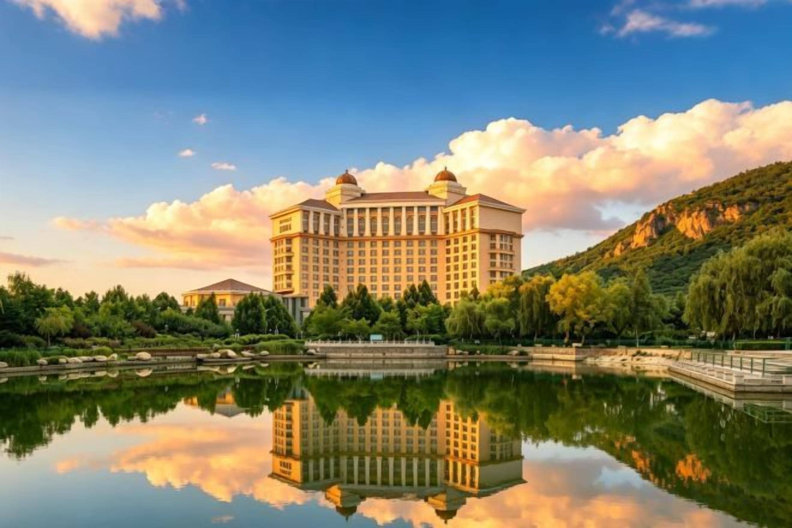 Un hotel grande con su reflejo en aguas tranquilas, rodeado de vegetación y un paisaje montañoso.