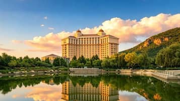 A large hotel with a reflection in a calm body of water, surrounded by greenery and a hilly landscape.