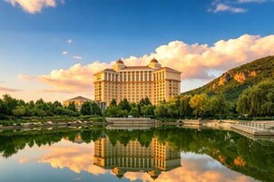 A large hotel with a reflection in a calm body of water, surrounded by greenery and a hilly landscape.