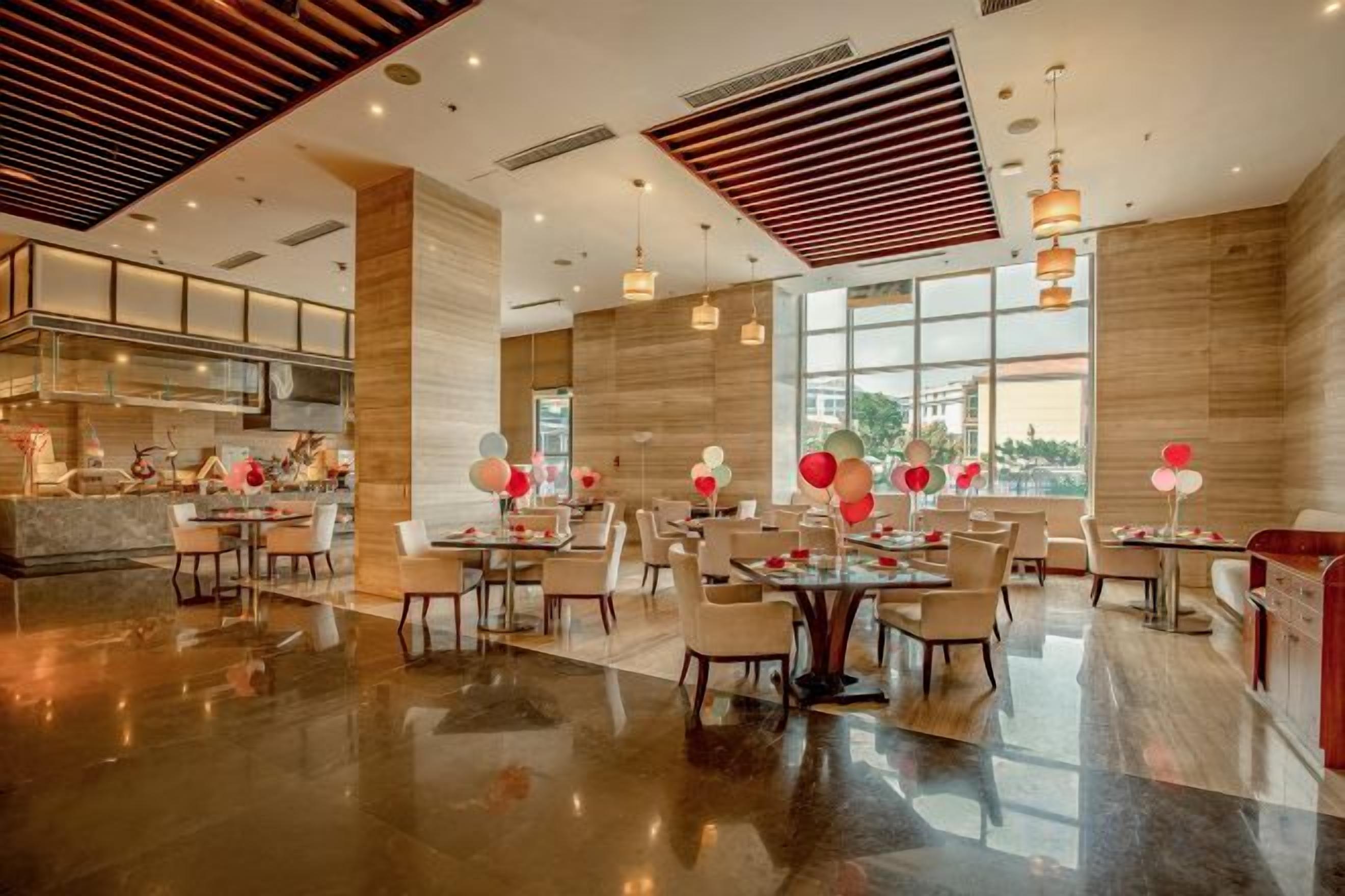 A spacious hotel lobby with multiple dining areas, a kitchen visible in the background, and large windows offering views.