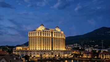 Un hotel grande de varios pisos con fachada simétrica, situado cerca de una masa de agua y con una montaña al fondo.