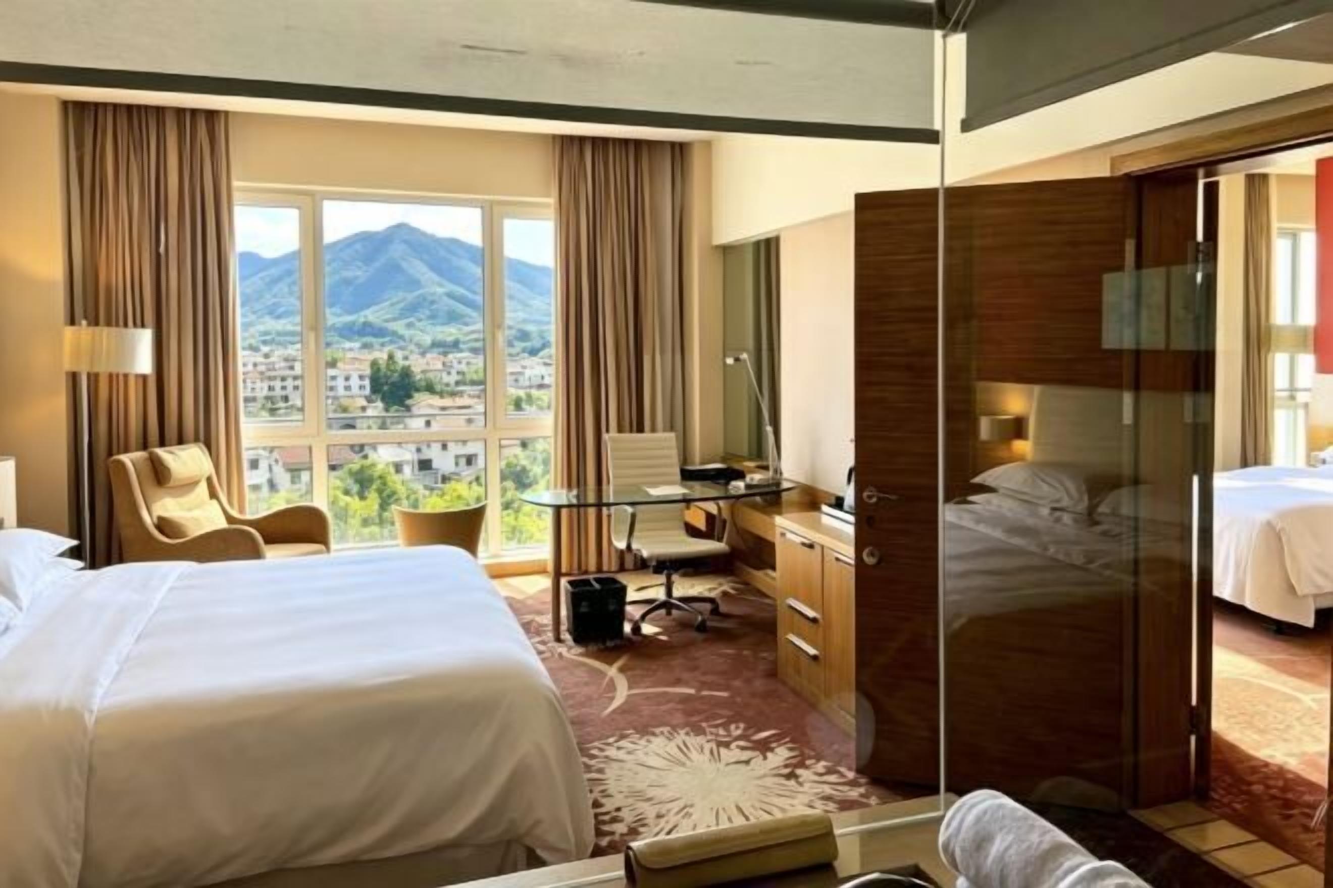 A hotel room with two beds, a desk, a chair, a desk lamp, a window with a mountain view, and a sliding door leading to another room.