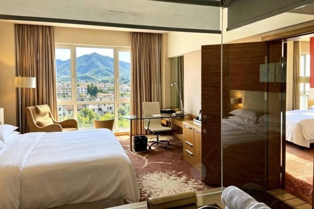 Hotelkamer met twee bedden, een bureau, een stoel, een bureaulamp, een raam met uitzicht op een berg en een schuifdeur naar een andere kamer.
