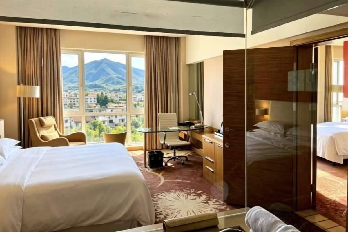 A hotel room with two beds, a desk, a chair, a desk lamp, a window with a mountain view, and a sliding door leading to another room.