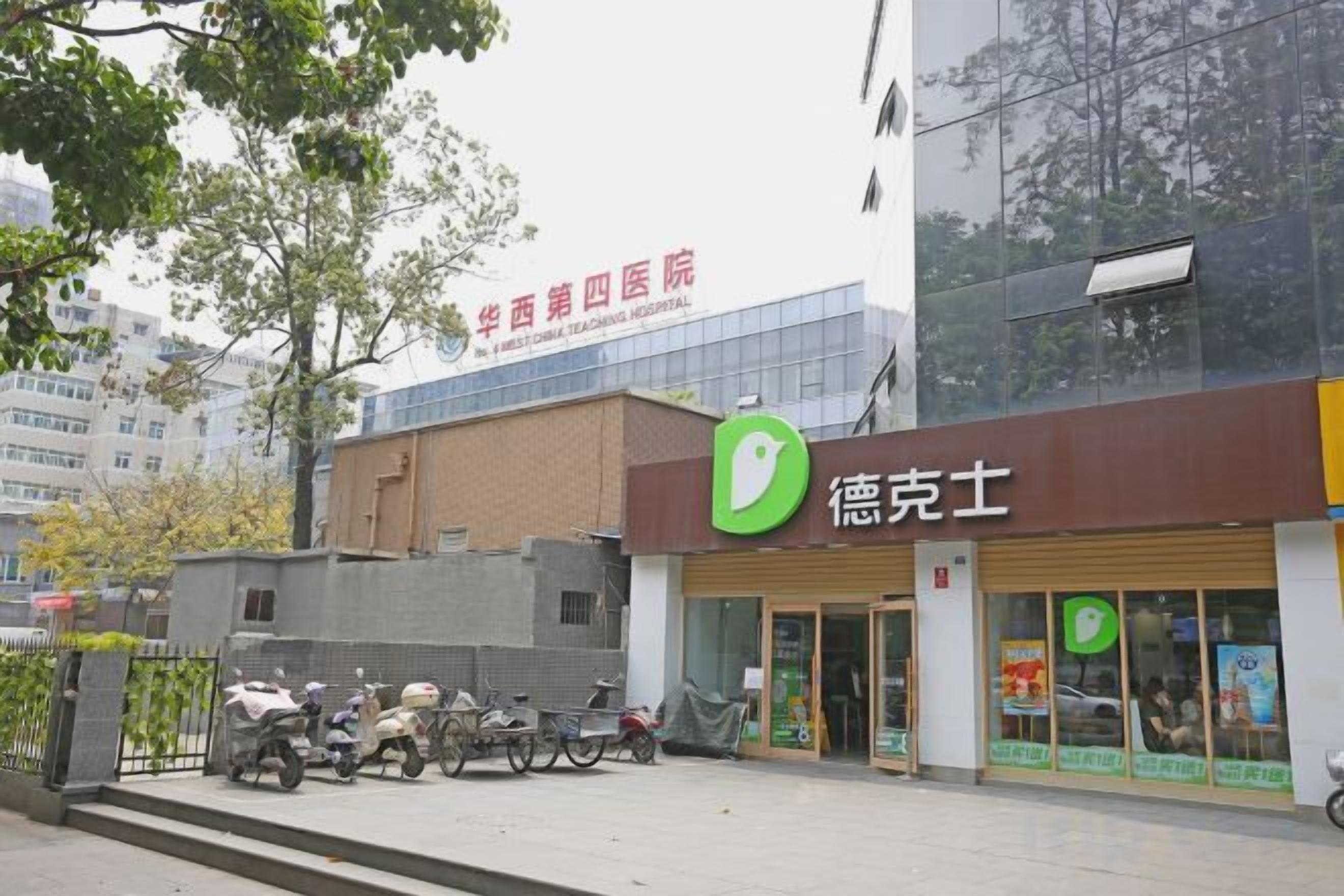 Een gebouw met een bord waarop '华西四医院' staat en een ander bord met een groen logo en tekst. Buiten staan scooters en een fiets geparkeerd.