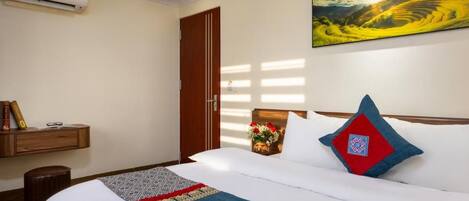 A hotel room with a large bed, a wooden nightstand, a wall-mounted air conditioner, a door, and a painting of a landscape.