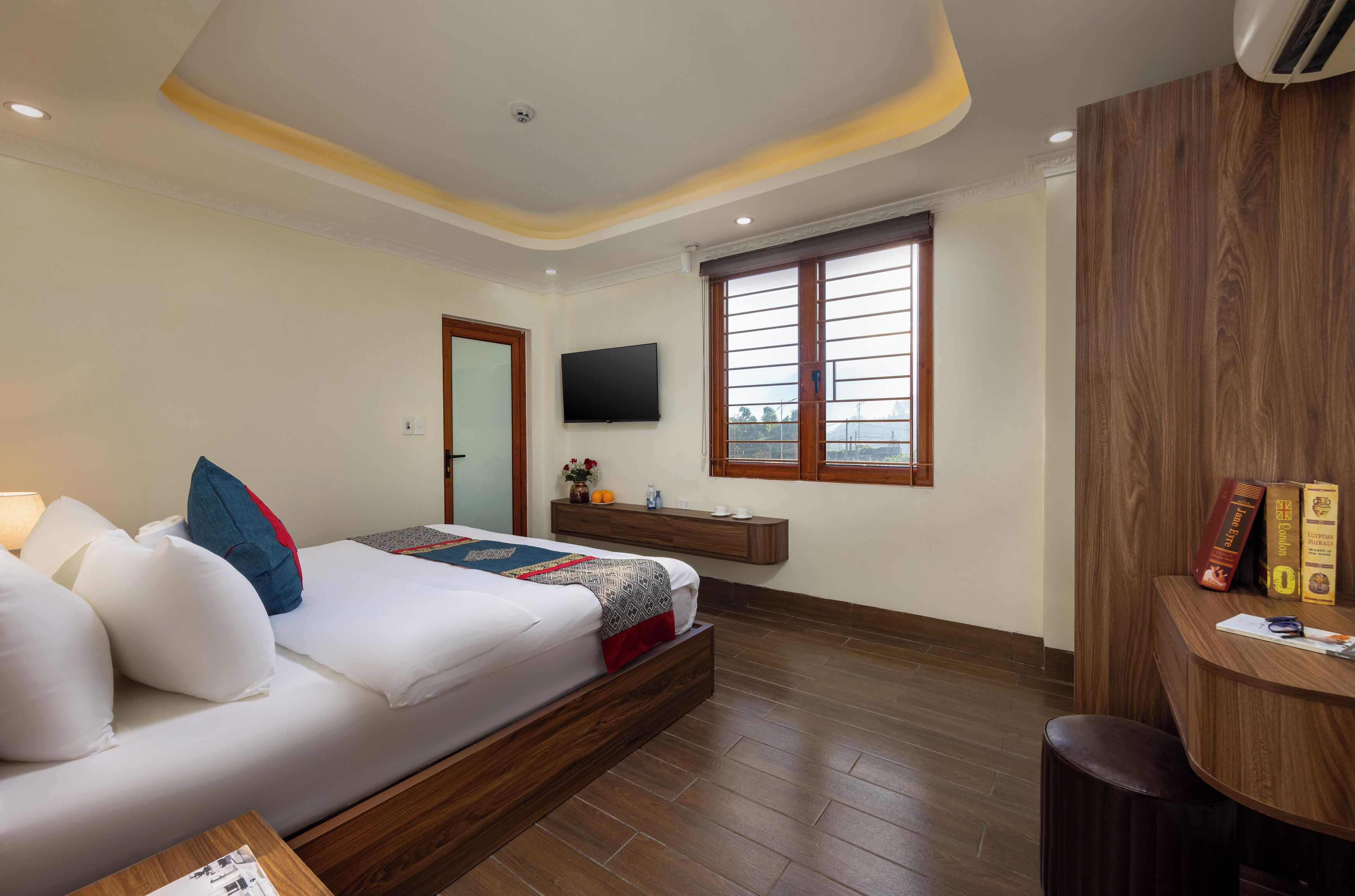 A modern bedroom with a bed, a TV mounted on the wall, a window with blinds, and a wooden desk with books and a lamp.