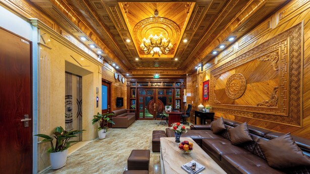 Un lobby lujoso con piso de mármol, una zona de estar central con sofá y otomanas, una araña y una pared decorativa con diseños intrincados.