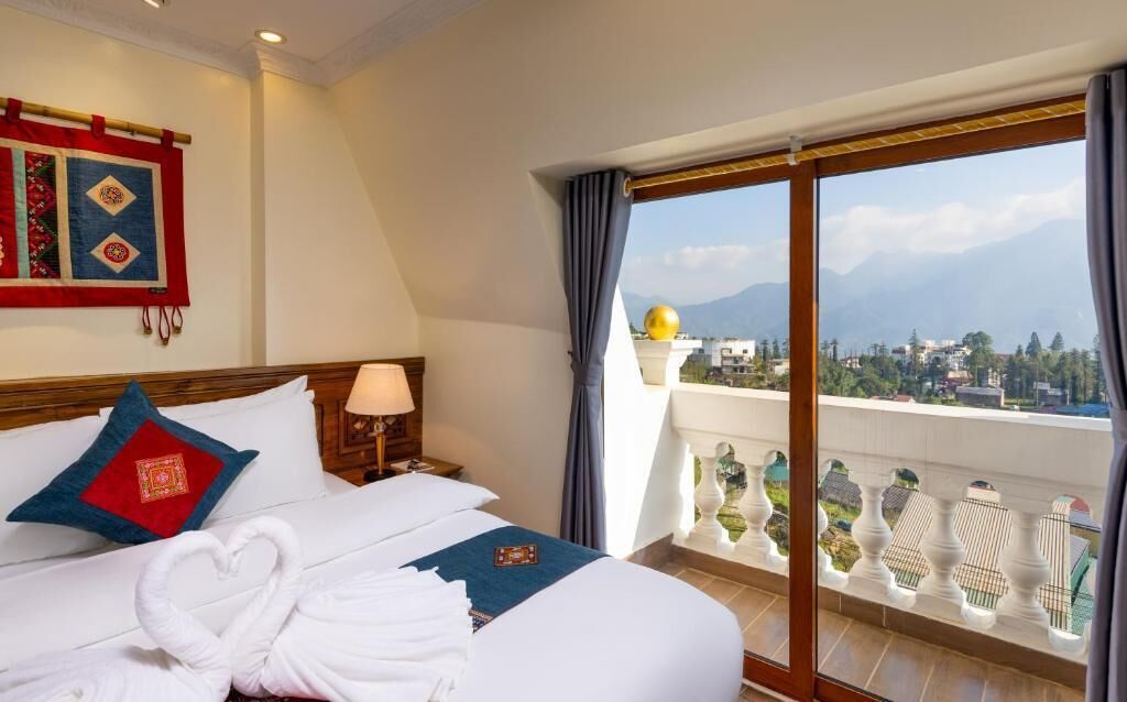 Una habitación de hotel con una cama, una mesita de noche, una lámpara, vistas a la ciudad y a las montañas a través de amplios ventanales, y un balcón con adornos.