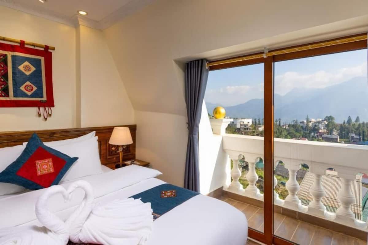 Una habitación de hotel con una cama, una mesita de noche, una lámpara, vistas a la ciudad y a las montañas a través de amplios ventanales, y un balcón con adornos.