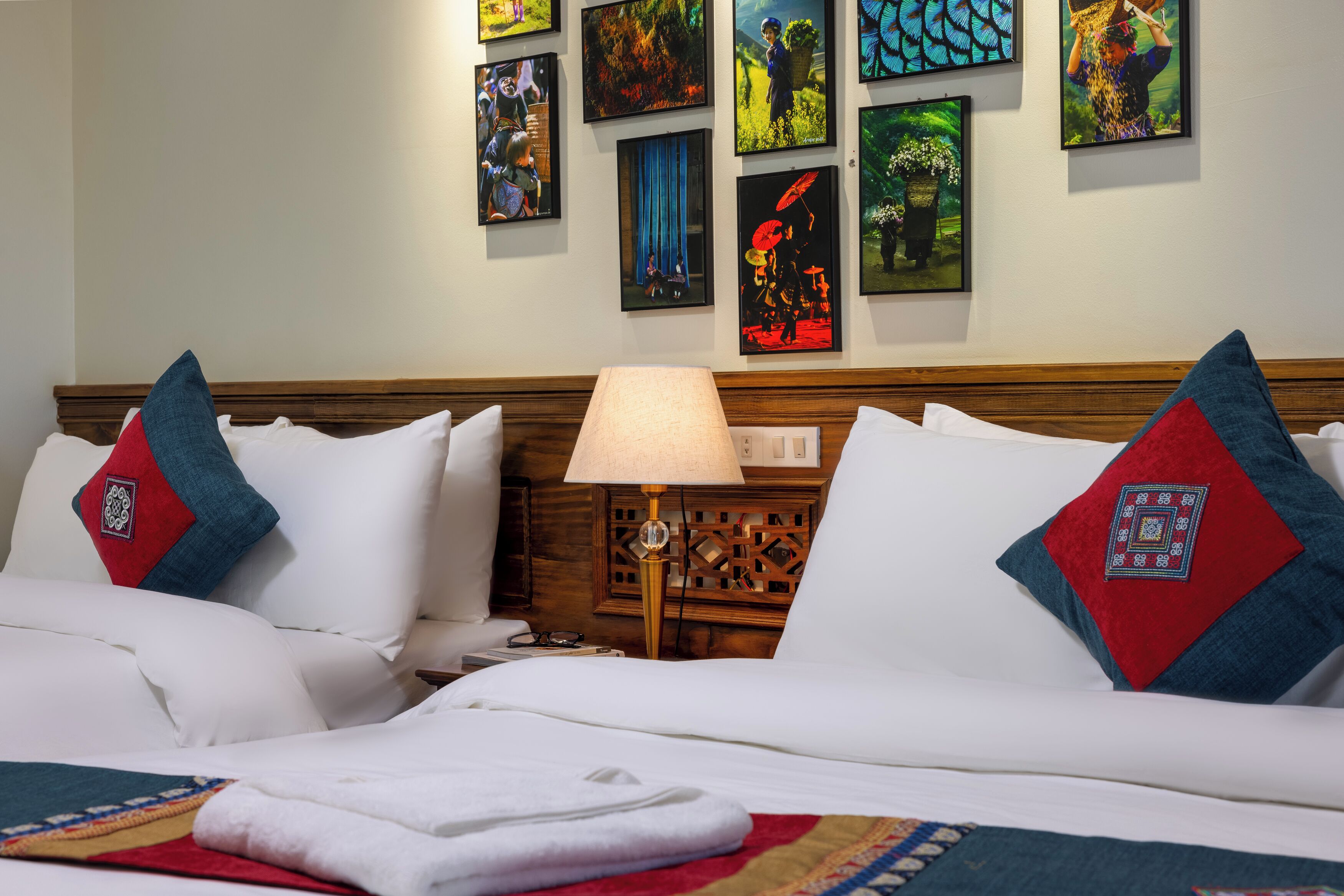Habitación de hotel con dos camas, cabecero de madera, mesita de noche con lámpara y una pared decorada con obras de arte enmarcadas.