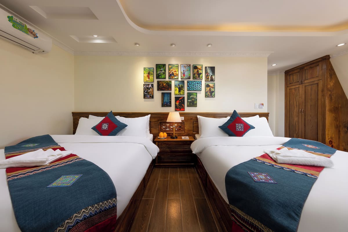 Una habitación de hotel con dos camas, una mesita de noche con lámpara, una pared con cuadros enmarcados y un armario de madera.