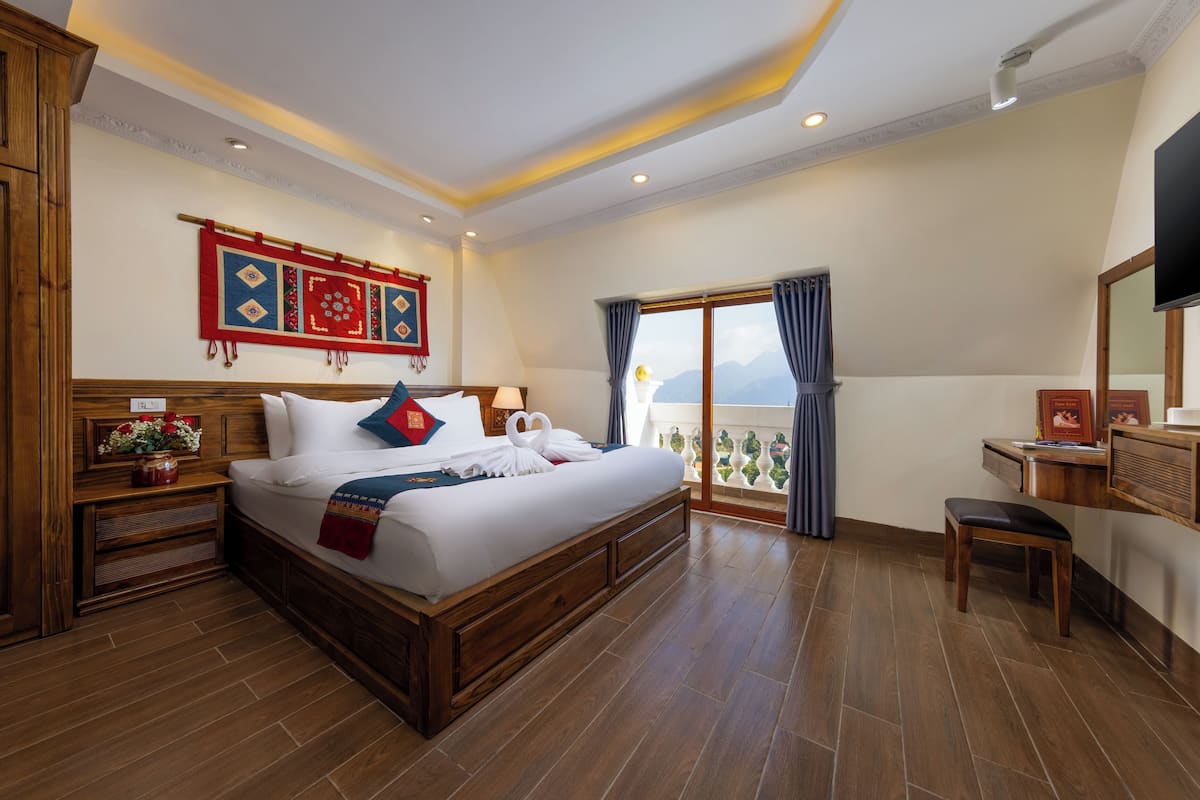 Amplia habitación de hotel con una cama grande, cabecera de madera, un escritorio y un balcón con vistas.