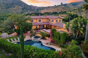 Une grande maison avec une piscine, entourée d’une végétation luxuriante et avec des collines en arrière-plan.