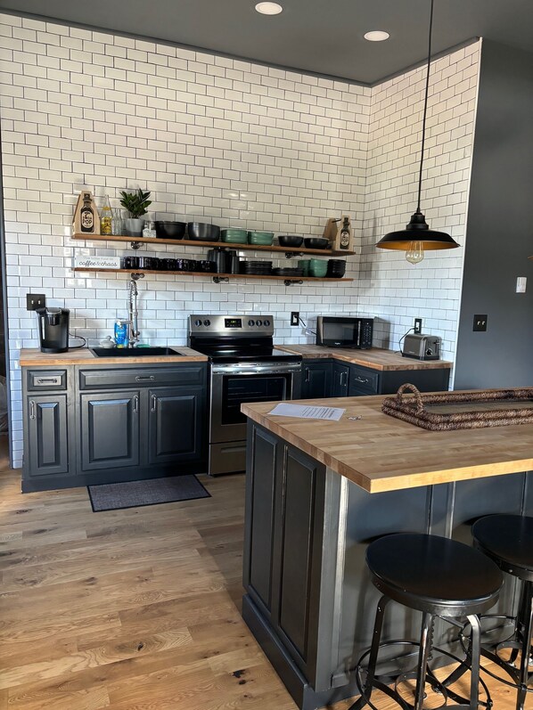 A modern kitchen with white brick backsplash, wooden countertops, and black cabinetry. There are shelves above the sink holding kitchenware, and a hanging light fixture above the island. - 2 Bedroom Restored Historic Landmark-Old Industrial Soda Factory in Mount Carmel (Mount Carmel)