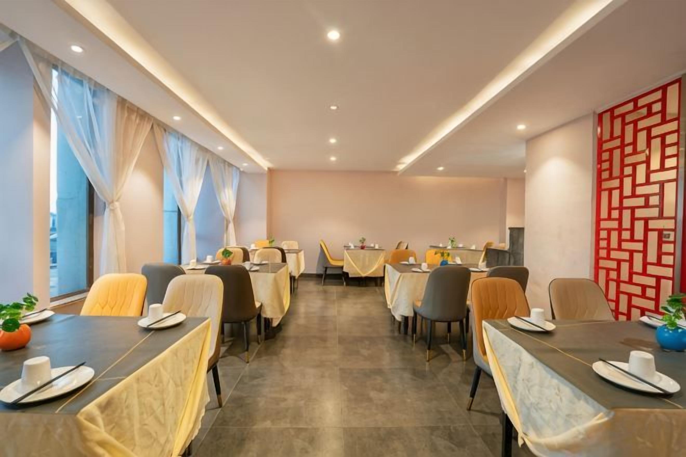 Un comedor de hotel con mesas preparadas para los huéspedes, ventanas amplias con cortinas y un diseño decorativo en la pared.
