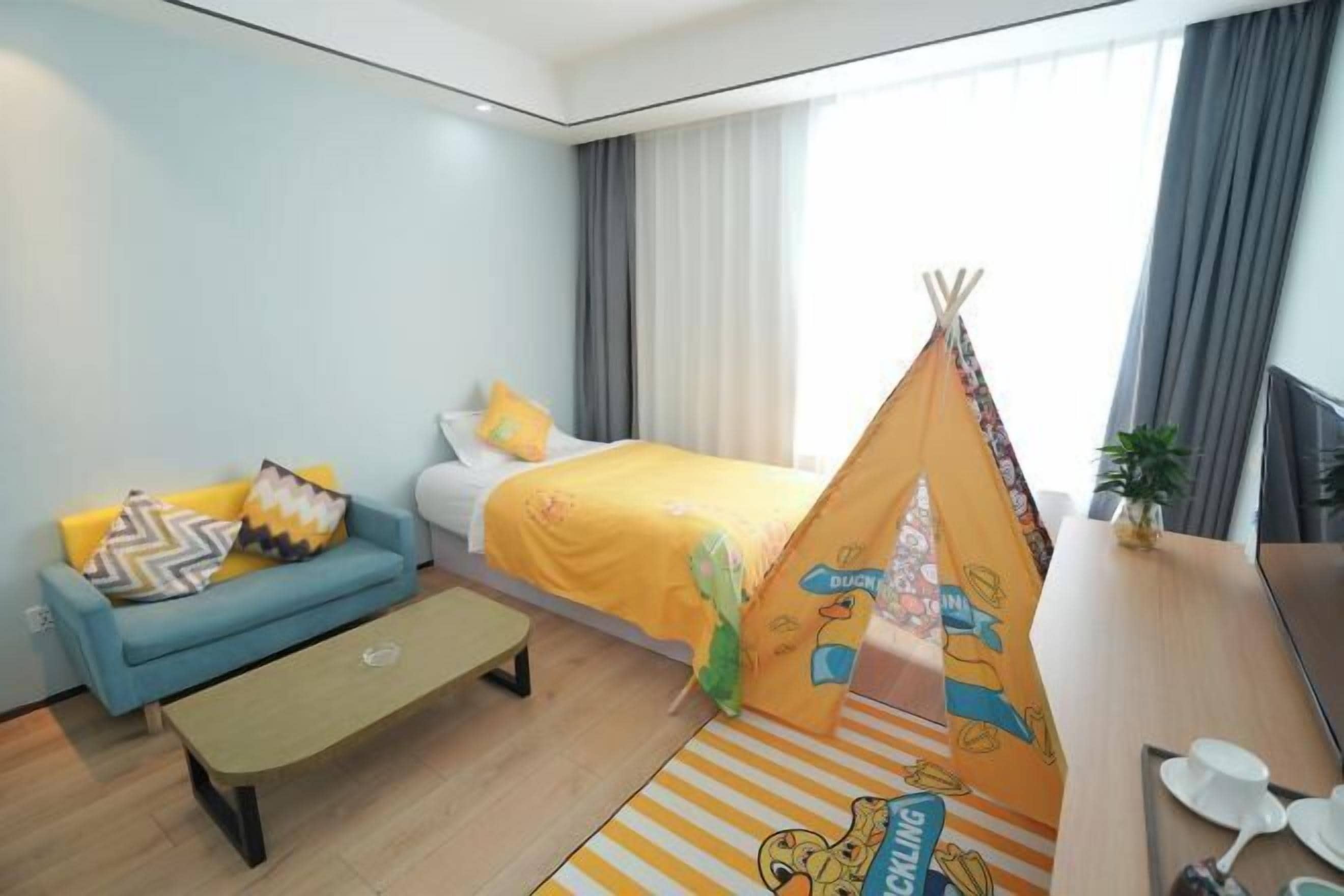 A hotel room with a bed, a blue sofa, a small table, a teepee, and a window with curtains.