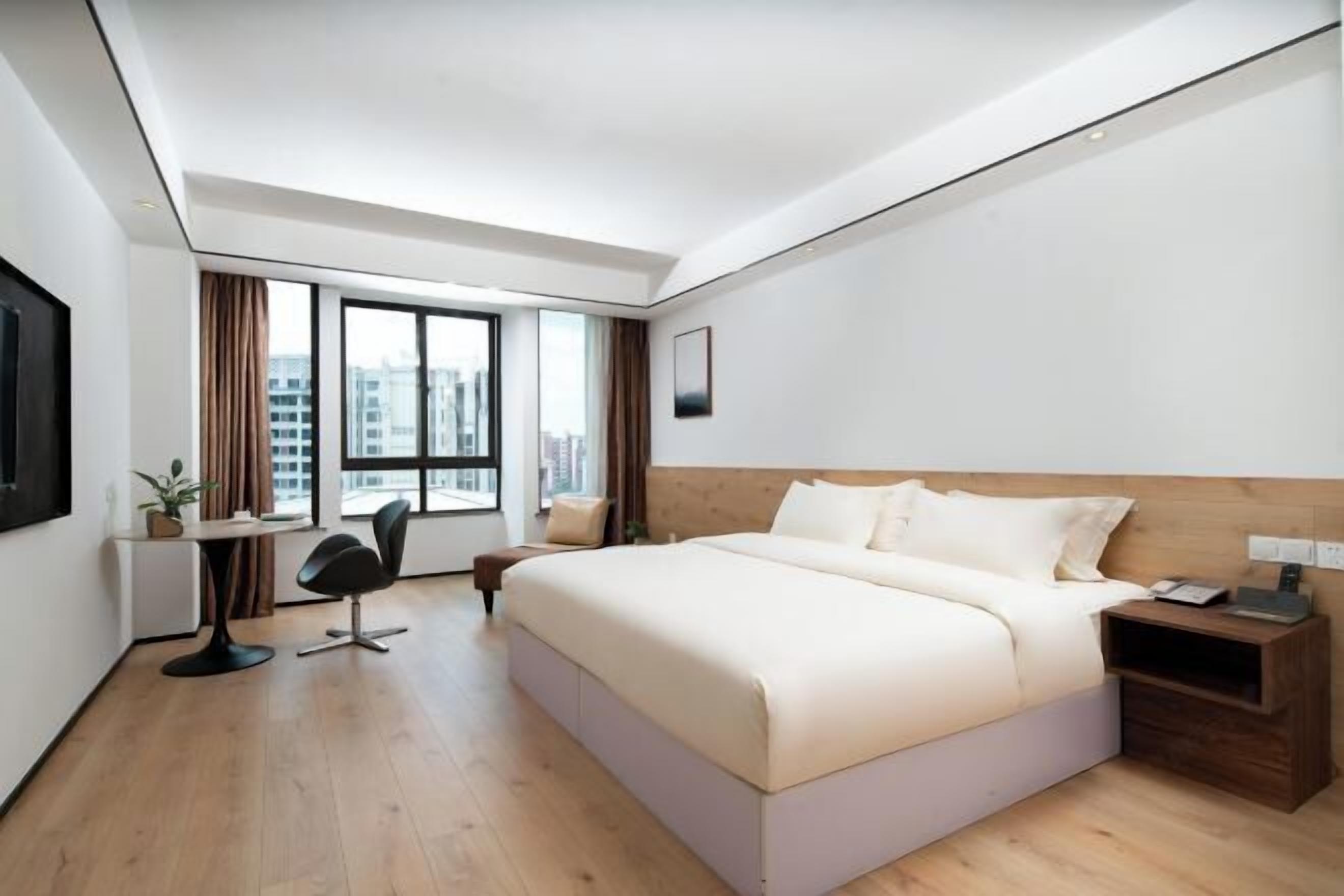 A modern hotel room with a large bed, a desk, a chair, and a view of the city.