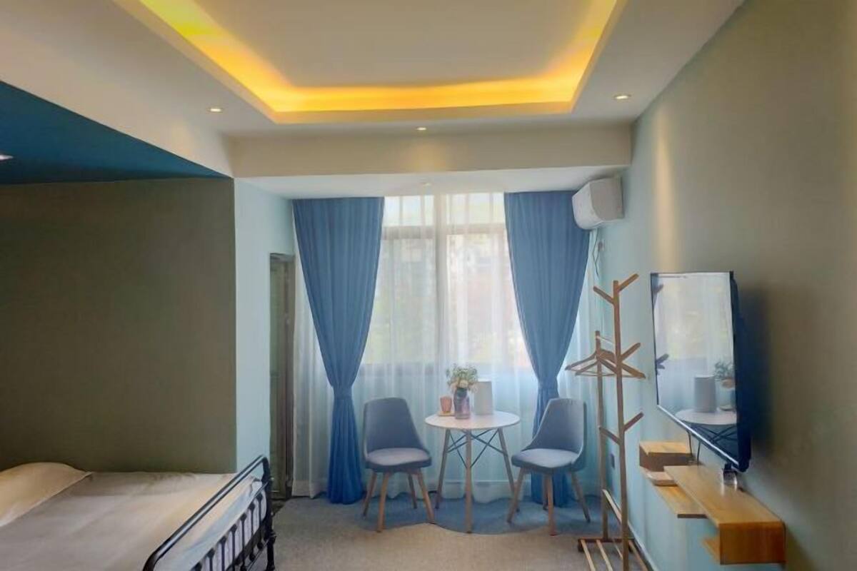 A hotel room with a bed, two chairs, a small table, a coat rack, a TV, and a window with curtains.