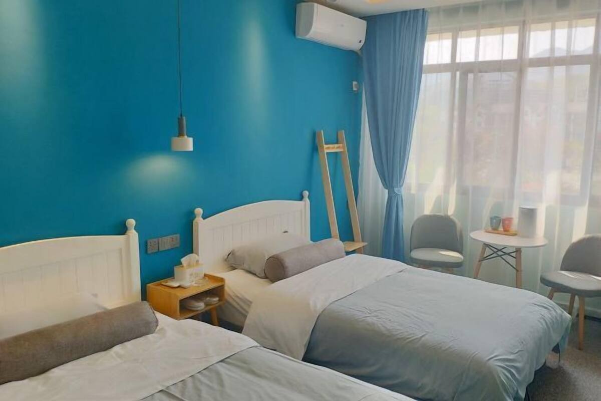 A hotel room with two beds, a bedside table with a lamp, a small table with chairs, and a window with curtains.