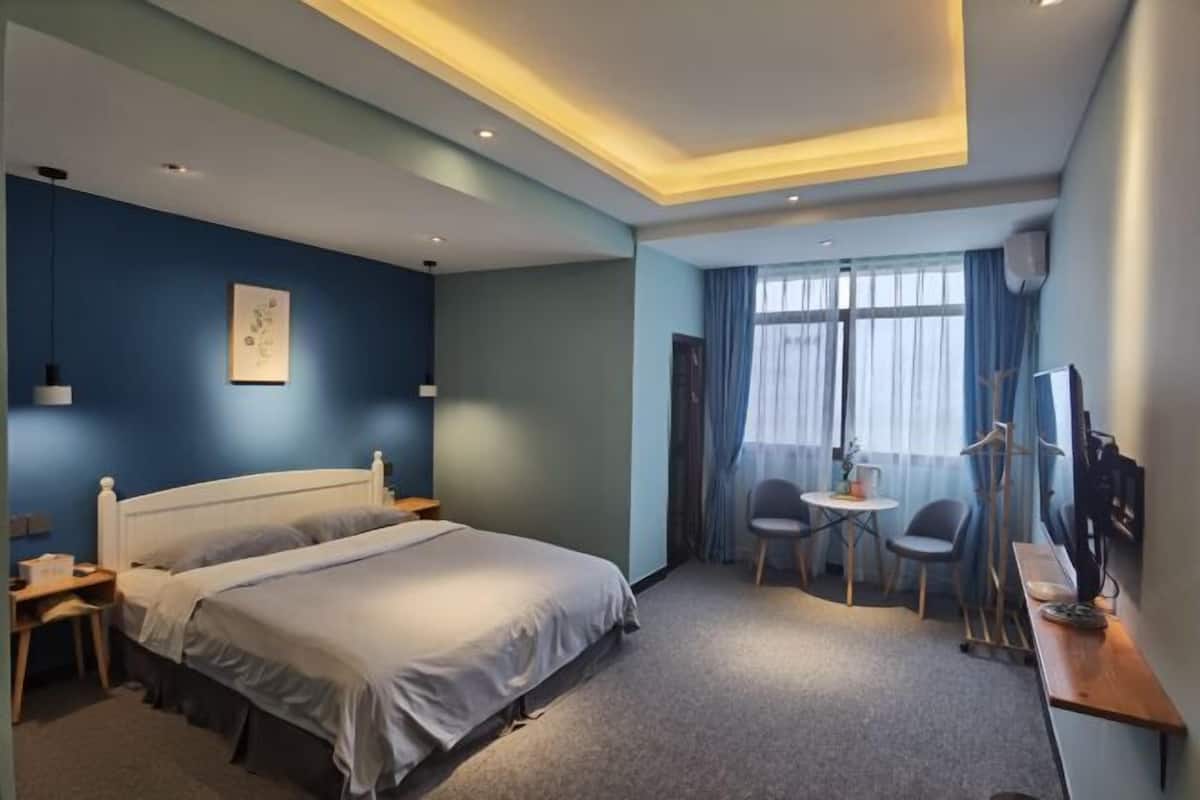 A hotel room with a bed, bedside tables, a desk, a chair, a small table, a TV, and a window with curtains.
