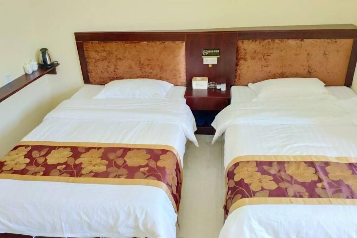 A hotel room with two beds, a wooden headboard, a nightstand with a clock, and a small shelf with items.