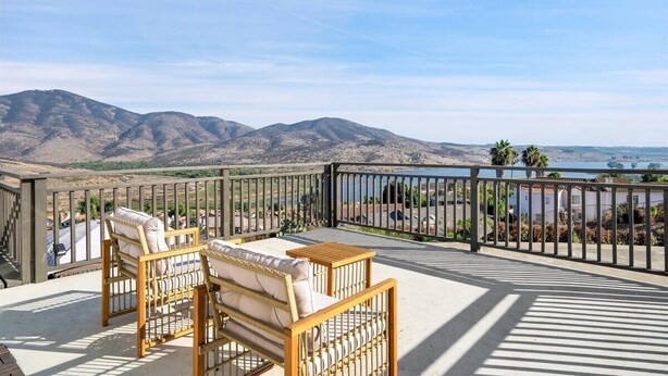 Une terrasse dotée de deux fauteuils en osier et d’une petite table, offrant une vue sur un paysage montagneux et une étendue d’eau.