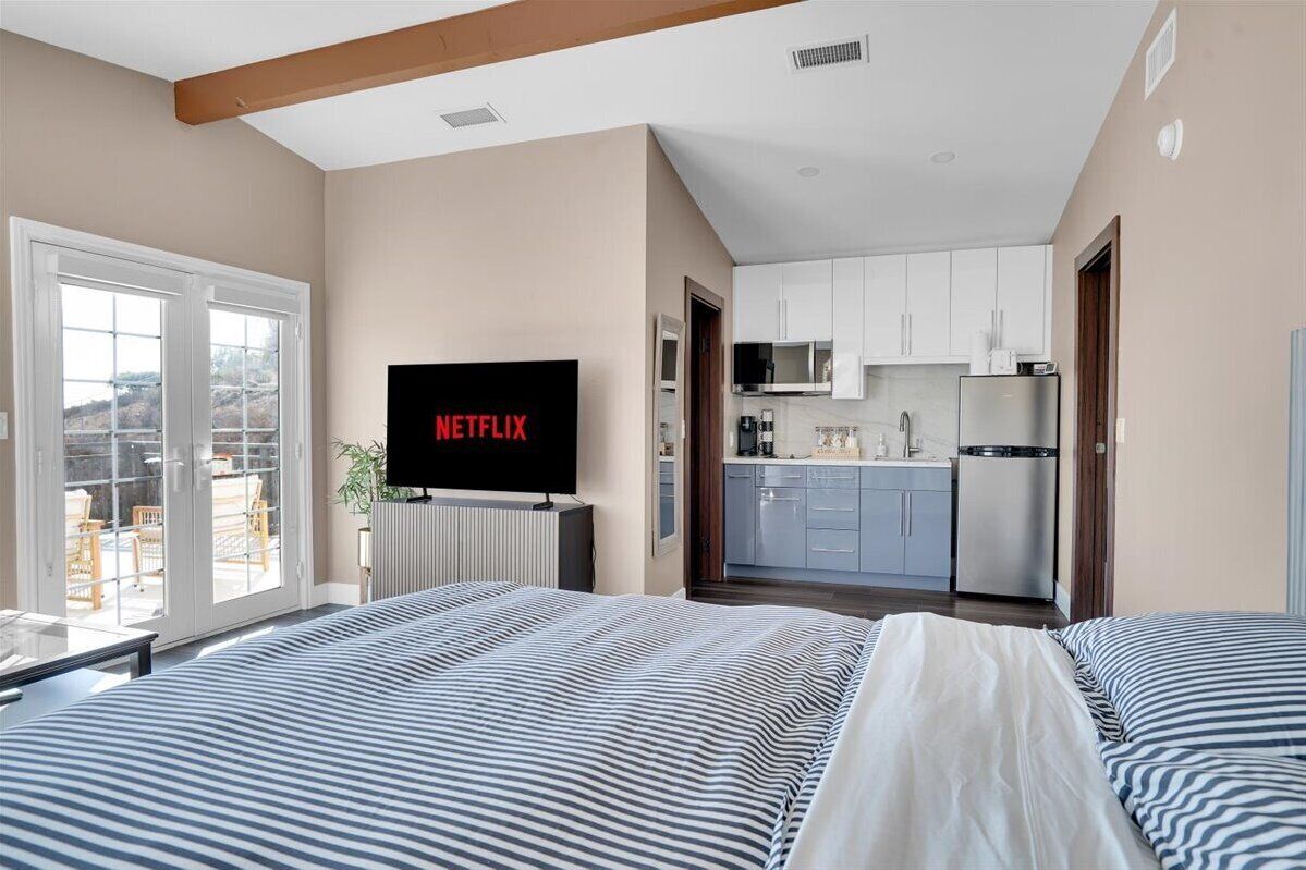 Habitació d'hotel moderna amb un llit gran, una televisió que mostra 'NETFLIX', una cuina americana i una vista per una porta de vidre.