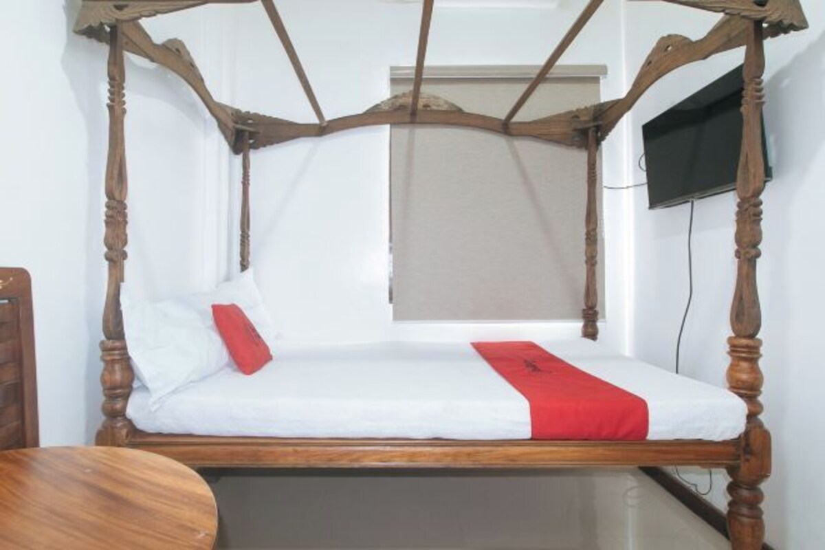 A single wooden bed with a white mattress and red accents, a television mounted on the wall, and a window with a blind.