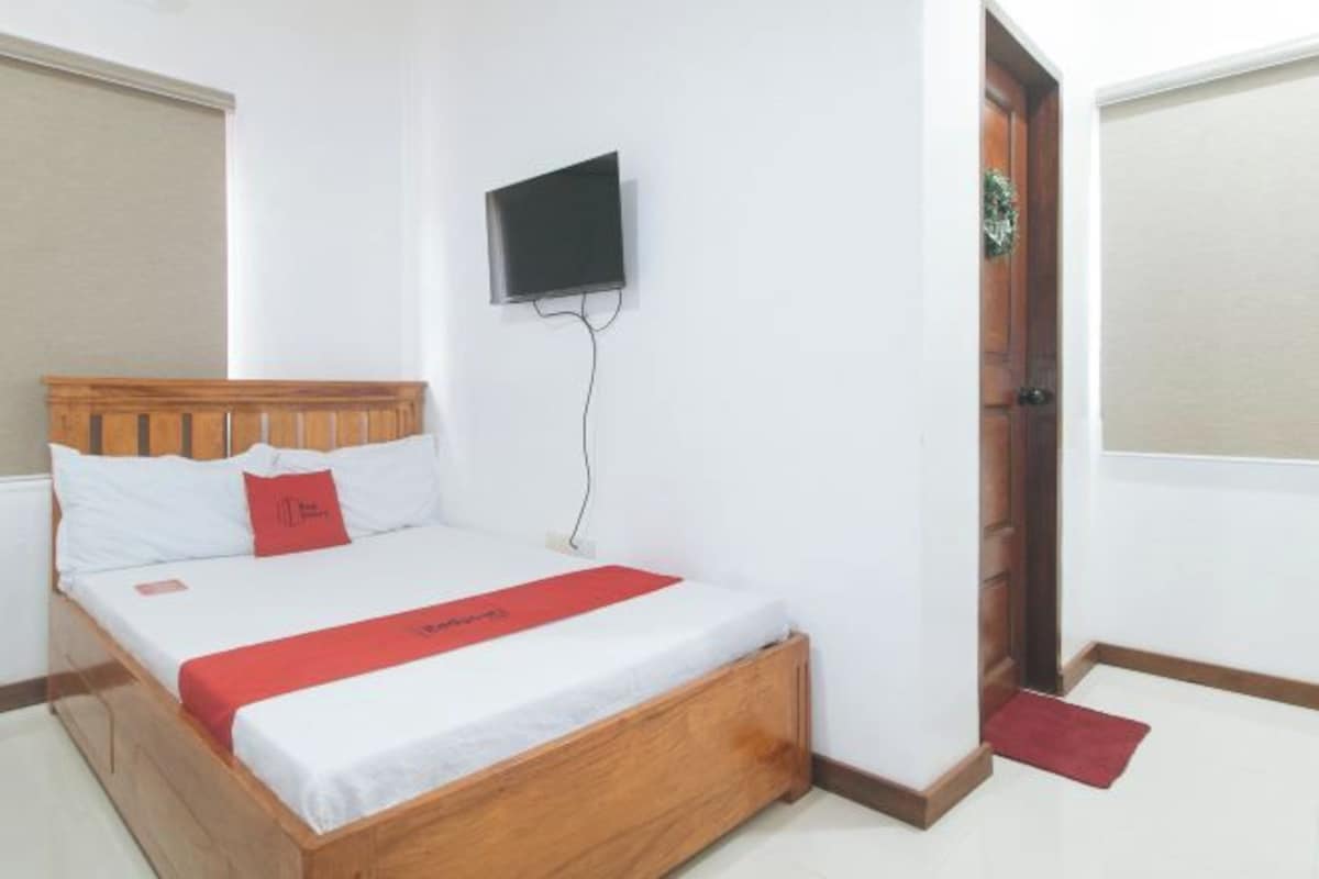 A neatly made bed with a wooden headboard, a flat-screen TV mounted on the wall, and a red doormat at the entrance.