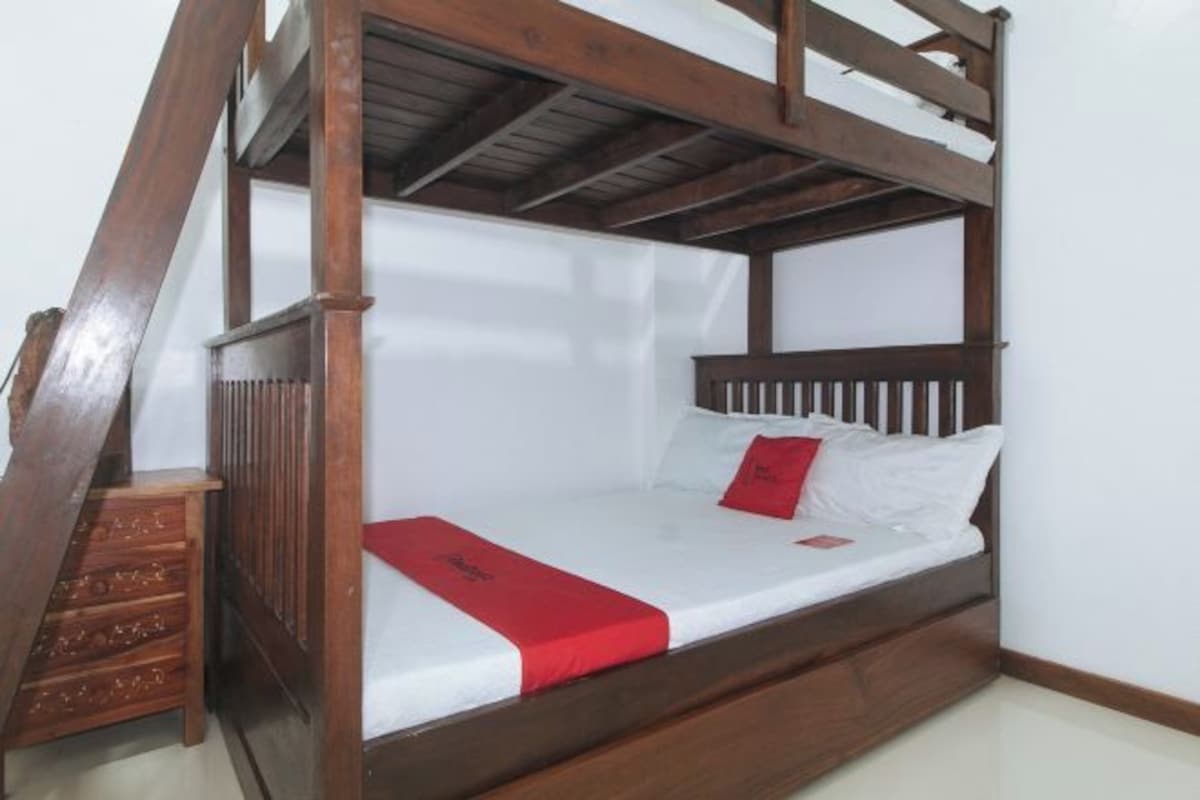 A wooden bunk bed with a red bedspread and pillows, a wooden ladder, and a small wooden chest of drawers.