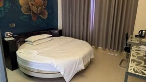 A round bed with a white bedspread, a nightstand with a lamp, a wall with floral wallpaper, and a window with curtains. - Guests City Daren Theme Hotel (Laibin)
