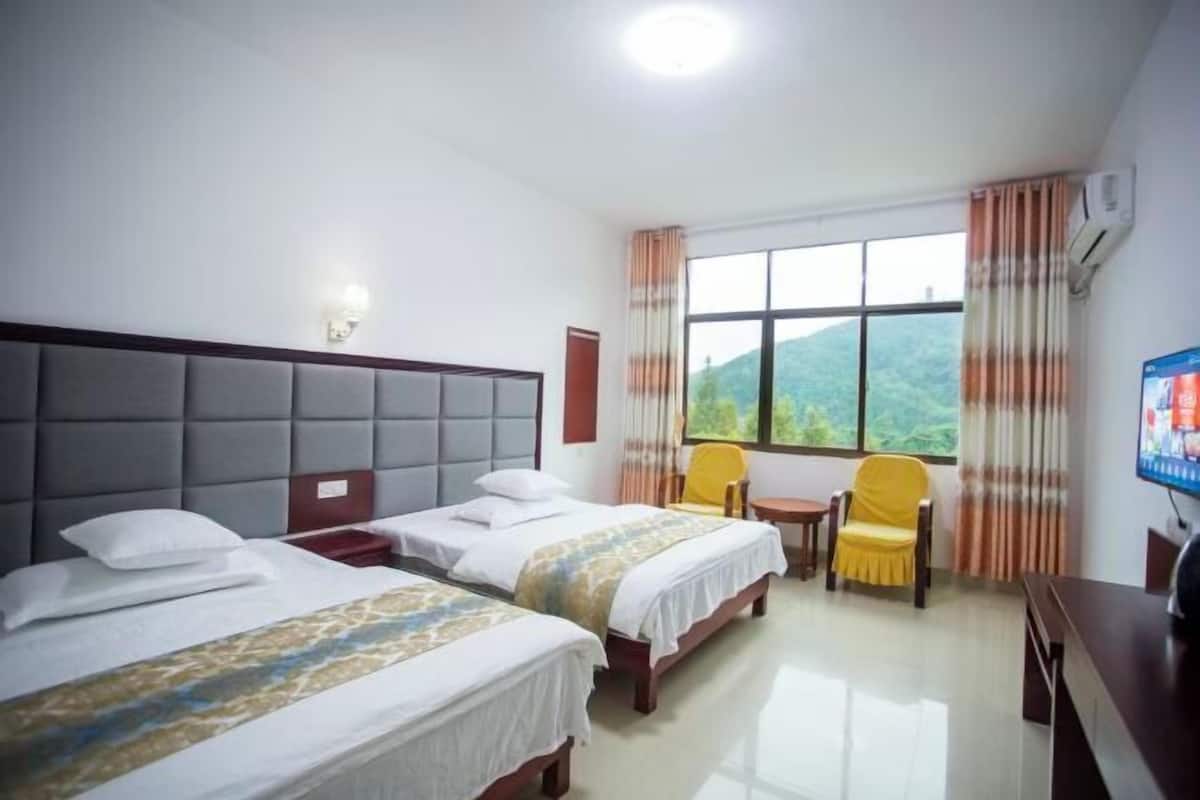 Ein Hotelzimmer mit zwei Betten, einem kleinen Tisch, einem gelben Sessel, einem Fernseher und einem Fenster mit Vorhängen.