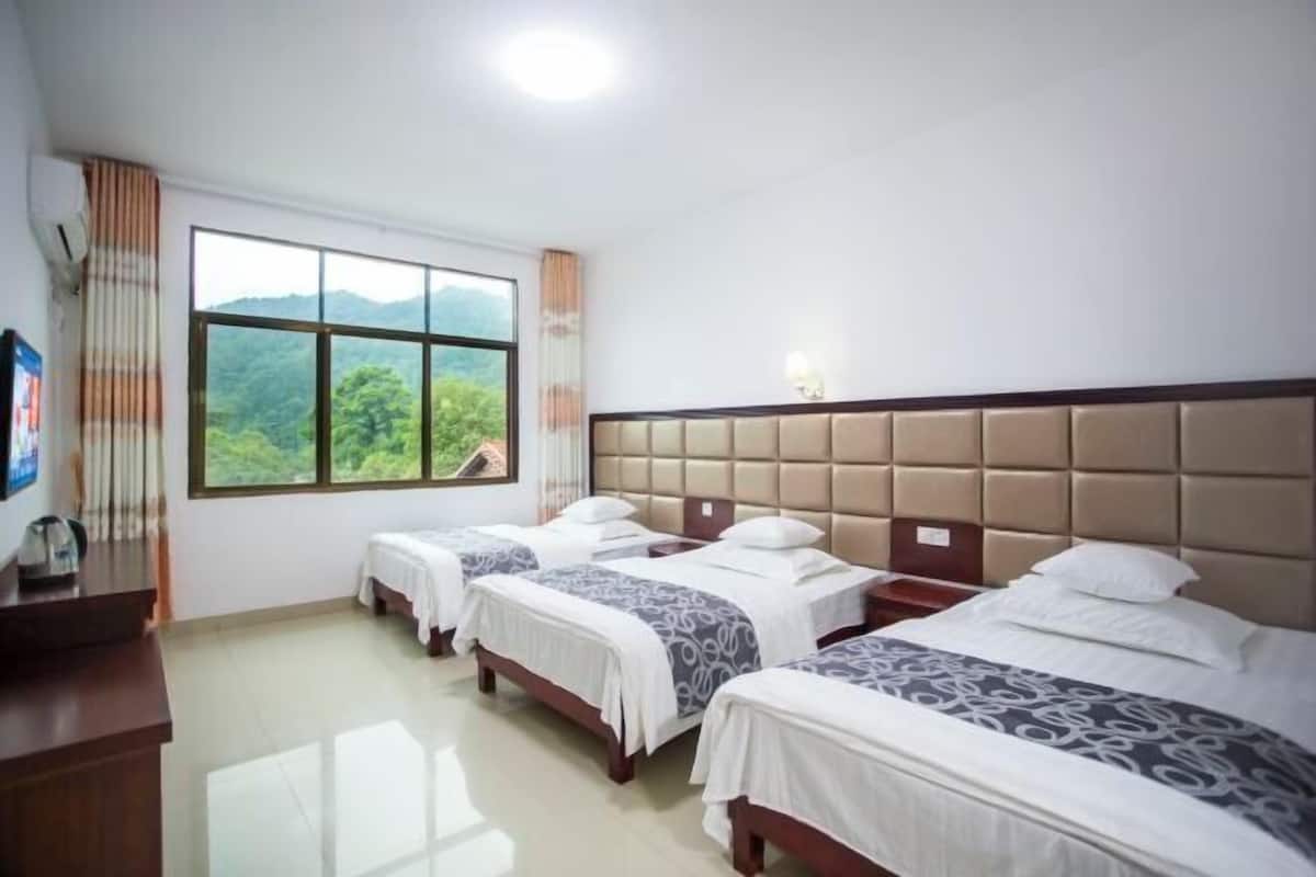 Ein Hotelzimmer mit drei Betten, einem Schreibtisch und einem großen Fenster mit Blick auf Grünflächen.
